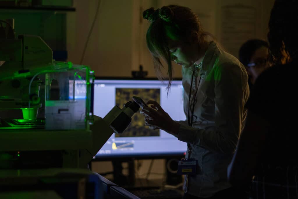 Dark microscopy room with student holding phone to the microscope lens to capture image. There is a computer screen showing confocal image in the background.