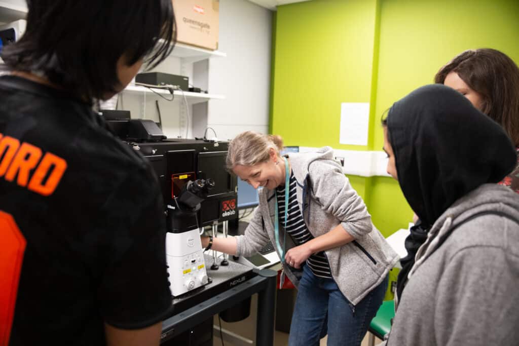 Three students look on as a researcher adjusts a piece of microscopy equipment