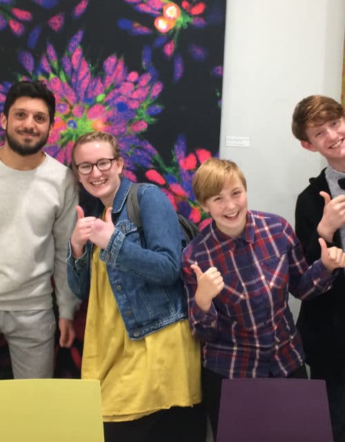 Five students smiling and showing thumbs up in front of large scientific artwork at the institute