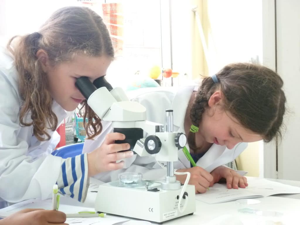 One girl looking through a microscope lens while the other takes notes
