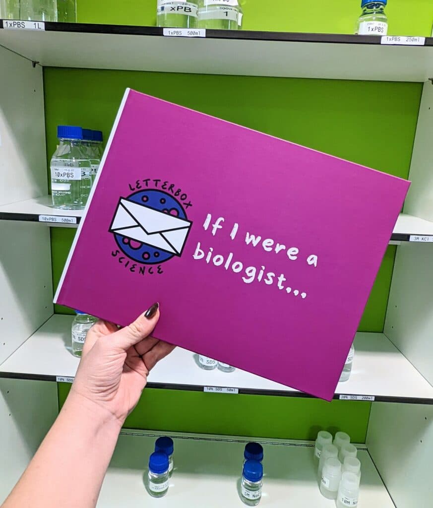A hand holding up a book with a purple cover showing the Letterbox Science logo and the text "If I were a biologist...". Behind is a green wall with white shelves stacked with lab glassware.