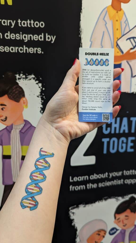 An outstretched arm with a colourful DNA helix tattoo in front of a Tattoo my Science banner and holding a small card with the tattoo design and some text