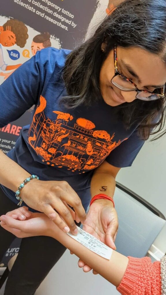 Woman wearing navy and orange Gurdon institute t-shirt applying a temporary tattoo on to the forearm of a member of the public