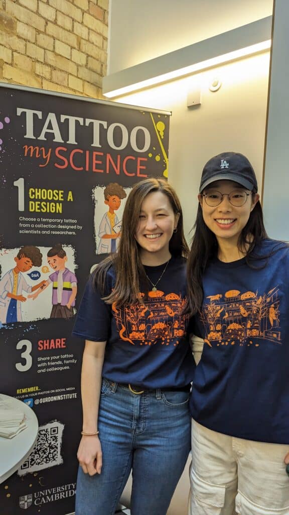 Two women standing in front of a Tattoo my Science banner, smiling and wearing navy and orange Gurdon Institute t-shirts