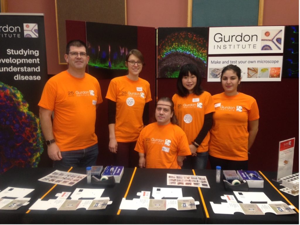 Five researchers at a public engagement event wearing orange Gurdon Institute t-shirts